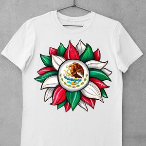 以下が含まれることがあります： 緑、白、赤の花のデザインが施された白いTシャツ。花の中心には、サボテンにとまっているワシが描かれたメキシコの紋章があります。
