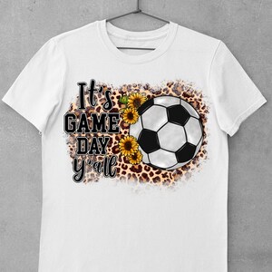 Puede incluir: Camiseta blanca con un bal&oacute;n de f&uacute;tbol blanco y negro, girasoles y un fondo de estampado de leopardo. El texto "It's Game Day y'all" est&aacute; impreso en la camiseta.