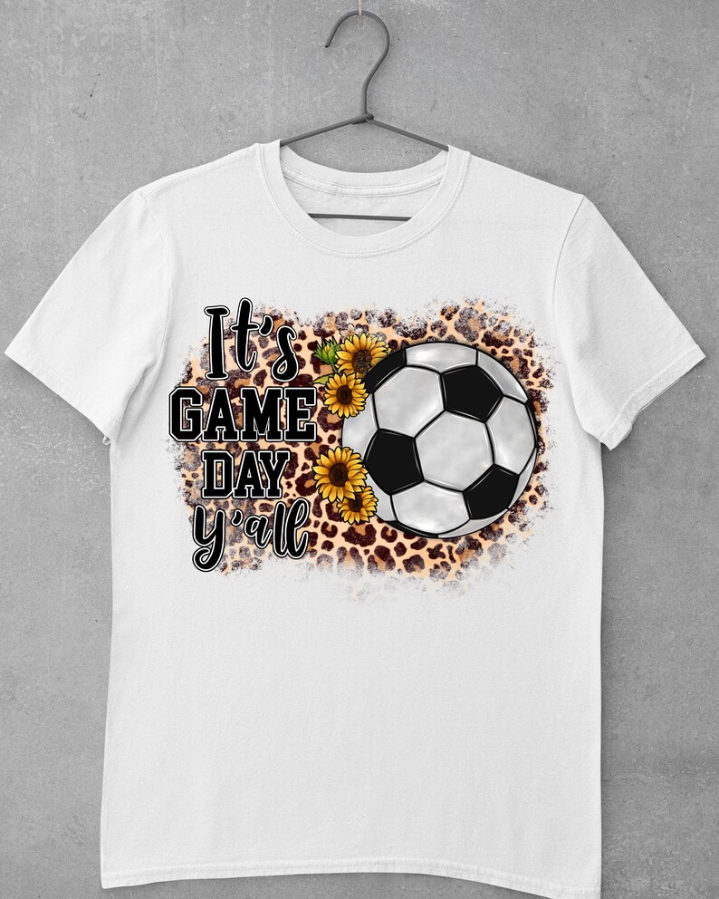 Puede incluir: Camiseta blanca con un bal&oacute;n de f&uacute;tbol blanco y negro, girasoles y un fondo de estampado de leopardo. El texto "It's Game Day y'all" est&aacute; impreso en la camiseta.