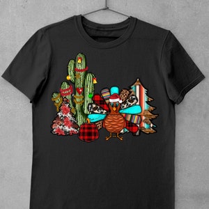 May include: A black t-shirt featuring a festive design with a turkey wearing a Santa hat, surrounded by cacti, a Christmas tree, and a plaid pumpkin.