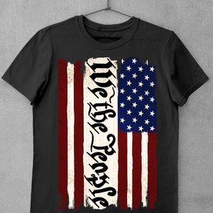 Peut inclure: T-shirt noir avec un motif de drapeau am&eacute;ricain us&eacute; et le texte "We the People" dans une police stylis&eacute;e.