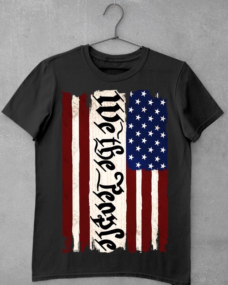 Peut inclure: T-shirt noir avec un motif de drapeau am&eacute;ricain us&eacute; et le texte "We the People" dans une police stylis&eacute;e.