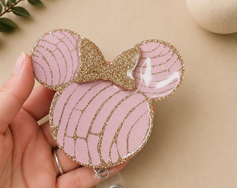 May include: A badge reel featuring a light pink concha bread shape with a gold glitter bow and gold glitter accents. The design includes a circular main shape with two smaller circular ears, mimicking a classic mouse silhouette, with a clear plastic clip.