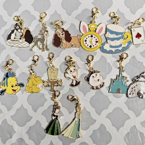 Puede incluir: Una colección de coloridos colgantes de esmalte con cierres dorados. Los colgantes presentan varios personajes de dibujos animados, incluyendo perros, una taza de té, un castillo y otros diseños caprichosos. Los colgantes están dispuestos sobre una superficie estampada.