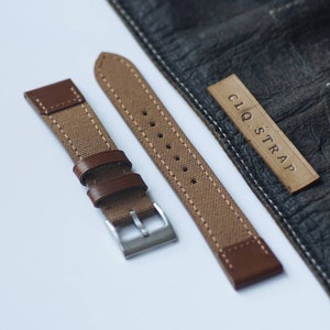 May include: Two brown canvas watch straps with brown leather accents and a silver buckle. The straps are laid out on a white surface. A brown leather patch with the text "CLQ. STRAP" is visible in the background.