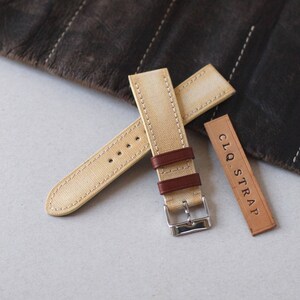 May include: A beige canvas watch strap with brown leather accents and a silver buckle. The strap is stitched with white thread and includes a small leather tag with the text "CLQ. STRAP".