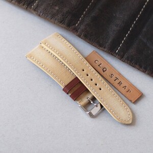 May include: A tan canvas watch strap with brown leather accents and white stitching. The strap has a silver buckle and is accompanied by a small leather tag that reads "CLQ. STRAP". The background includes a rolled-up brown leather item.