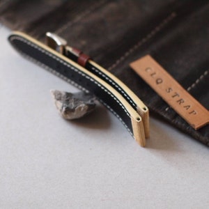 May include: A close-up of a black and beige watch strap with white stitching. The strap has a silver buckle and a brown leather accent. A small, light-colored rock is next to the strap. A leather tag with the text "CLQ STRAP" is also visible.