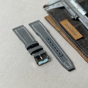 May include: Two grey watch straps with white stitching and silver buckles. The straps are made of a textured fabric. A leather tag with the text "CLOSTRAP" is visible. The straps are displayed on a light grey surface.