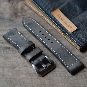 Op de afbeelding: Een grijs leren horlogebandje met witte stiksels en een zilveren gesp. Het bandje ligt op een houten oppervlak en heeft het woord "CLOSTRAP" gedrukt op een bruin leren label.