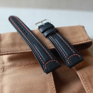May include: Two black leather watch straps with orange stitching. The straps are laid out on a brown canvas surface.