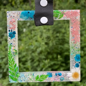 May include: A square-shaped clear resin picture frame with a white background and a variety of pressed flowers and greenery. The frame has a black strap with white buttons.