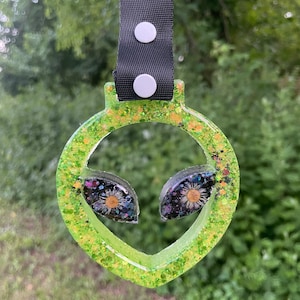 May include: Green alien face keychain with glitter and black and white flower accents. The keychain has a black strap with white buttons.