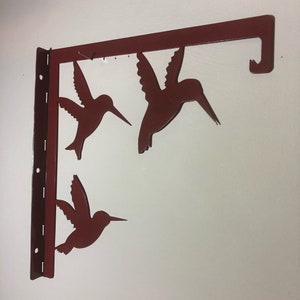 May include: A metal wall-mounted shelf with three hummingbird silhouettes cut out. The shelf is painted a deep red color and has a hook on the right side.