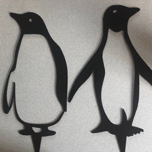 May include: Two black metal penguin silhouettes, standing upright on a white background. The penguins are facing different directions.