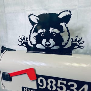 May include: Black metal raccoon mailbox topper with a playful expression, peeking over the top of a white mailbox with a red flag and the number 98534.