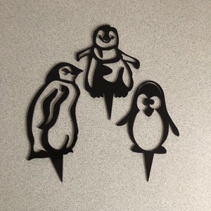 May include: Three black penguin cake toppers. The penguins are in different poses, one is standing upright, one is standing with its wings out, and one is standing with its head tilted to the side.