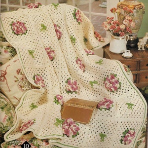 Könnte beinhalten: Eine gehäkelte Tagesdecke mit weißem Hintergrund und grünen und rosa floralen Akzenten. Die Decke liegt über einem Stuhl mit einem floral gemusterten Stoff.