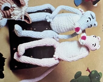 May include: Two crocheted cat toys, one light blue and one pink, with black eyes and pink noses. The toys are sitting on a black surface.