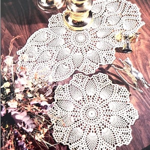 Könnte beinhalten: Zwei weiße Häkeldeckchen, eines groß und eines klein, mit einem zarten, durchbrochenen Muster. Die Deckchen liegen auf einem Holztisch mit einem goldenen Kerzenleuchter und anderen Dekorationsgegenständen.