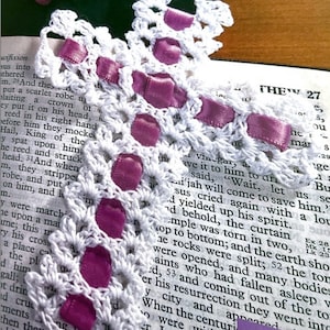 May include: A white crocheted bookmark with a purple ribbon woven through it. The bookmark is placed on a page of a book with the text "THEW 27" visible.