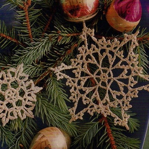 May include: Two crocheted snowflakes, one white and one beige, are displayed on a green pine branch with red and purple ornaments.