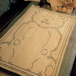 May include: A crocheted baby blanket with a bear design in a cream color. The blanket is on a bed with three teddy bears.