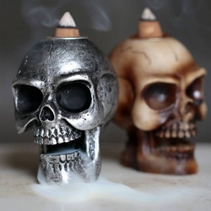 May include: Two ceramic skull incense holders, one silver and one brown, with incense cones on top. Smoke is coming from the silver skull.