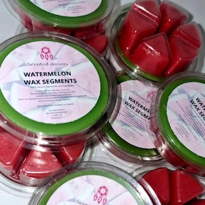 Könnte beinhalten: Mehrere transparente Plastikbehälter gefüllt mit roten, wassermelonenförmigen Wachssegmenten. Die Behälter haben grüne Ränder und Etiketten mit der Aufschrift "Watermelon Wax Segments" mit dem Markennamen "Scented Dreams."