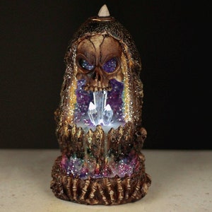 May include: A ceramic incense burner shaped like a skull with a hooded cloak. The skull has glowing purple eyes and a crystal inside. The base is decorated with purple and gold glitter.