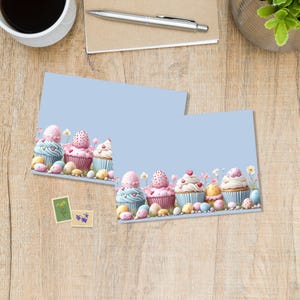 Può includere: Due biglietti azzurri con un design di cupcake. I cupcake hanno glassa rosa, bianca e blu, decorati con cuori e uova di Pasqua. I biglietti sono su una superficie di legno con una tazza di caffè, una penna e una pianta.