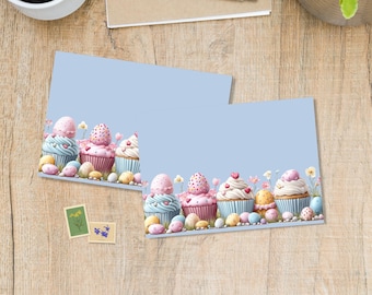 May include: Two light blue cards with a cupcake design. The cupcakes have pink, white, and blue frosting, decorated with hearts and Easter eggs. The cards are on a wooden surface with a coffee cup, pen, and plant.
