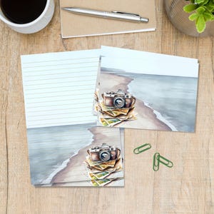 May include: Stationery set featuring a watercolour illustration of a vintage camera resting on a stack of photographs against a beach shoreline. The paper includes horizontal lines for writing and the design is repeated on a smaller rectangular card.