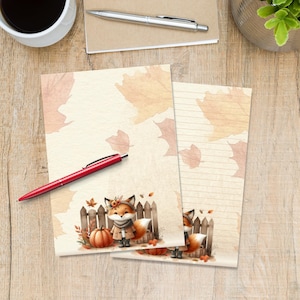 Può includere: Due fogli di carta da lettere con un'illustrazione ad acquerello di una volpe che indossa una sciarpa e un cappotto, in piedi davanti a una staccionata di legno con una zucca. Lo sfondo è beige chiaro con foglie autunnali arancioni sparse.