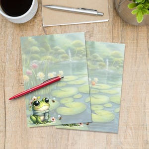 Op de afbeelding: Twee vellen briefpapier met een serene vijverscène met waterlelies, bloemen en een cartoonkikker met een sjaal en bloemenkrans. Een rode pen ligt op het briefpapier. Het briefpapier is gelijnd.