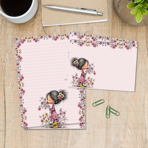 Può includere: Un set di cancelleria con un foglio di carta da lettera a righe e un biglietto vuoto, entrambi con sfondo rosa e bordo floreale. Le illustrazioni raffigurano una ragazza in abito rosa che tiene dei fiori. Il set è su una superficie di legno.