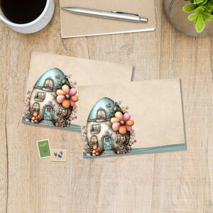 May include: Two blank stationery cards with a whimsical illustration of a fairy house with a blue roof and a large flower. The cards are decorated with floral accents and sit on a wooden surface. Two postage stamps are also visible.