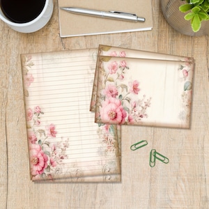 Op de afbeelding: Briefpapier set met een gelijnd notitieblok en twee briefkaarten. De set is versierd met een vintage bloemenontwerp in roze en crèmetinten. Het notitieblok heeft lijnen om op te schrijven en de kaarten zijn blanco. Groene paperclips zijn ook zichtbaar.