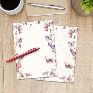 May include: Two sheets of stationery with floral and butterfly designs. The stationery has a cream background with watercolour illustrations of pink and purple flowers and butterflies. A red pen rests on the stationery. A cup of coffee is in the background.