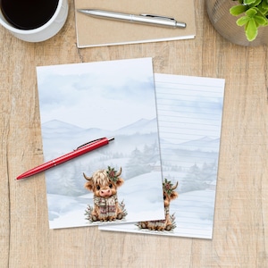 Op de afbeelding: Twee vellen briefpapier met een aquarelillustratie van een pluizige Schotse hooglander met een geruite sjaal en een takje hulst. De achtergrond toont een besneeuwd berglandschap met dennenbomen. Een rode pen ligt op het papier.