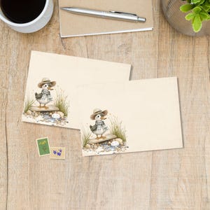 May include: Two cream-colored stationery cards with a charming illustration of a duck wearing a hat and scarf. The duck stands on a wooden plank with grass and stones. Two small stamps are also visible.
