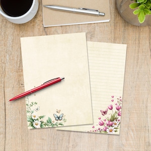 Può includere: Due fogli di carta da lettere con disegni floreali e farfalle. Un foglio è vuoto, l'altro a righe. Una penna rossa poggia sul foglio bianco, e una tazza di caffè e una penna sono sullo sfondo. La carta da lettere ha uno sfondo color crema.