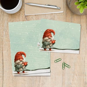 May include: Stationery set featuring a gnome illustration on lined paper and a matching card. The gnome wears a red hat, grey coat, and scarf, holding a steaming red mug against a light green background with white falling snow and white ground.