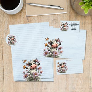 Pode incluir: Conjunto de papelaria com uma ilustração de vaca caprichosa rodeada por flores cor-de-rosa e borboletas. Inclui papel pautado, um autocolante, uma etiqueta de endereço e um cartão pequeno. A vaca tem um nariz rosa e está a sorrir.