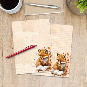 Pode incluir: Duas folhas de papel de carta com uma ilustração de raposa. A raposa é laranja e branca, sentada com um livro aberto, rodeada por folhas de outono. Uma caneta vermelha repousa sobre o papel de carta, com uma chávena de café e uma caneta no fundo.