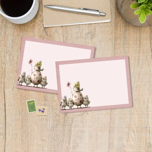 May include: Two pink stationery cards with a light pink border. One card features a group of cartoon chicks and an egg with pink spots. The other card is blank. The cards are on a wooden surface.