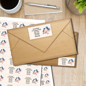 Può includere: Busta di carta kraft con etichette indirizzo raffiguranti uccelli blu su un ramo fiorito. Le etichette includono il nome "Sarah Black" e i dettagli dell'indirizzo. Sono visibili una tazza di caffè e una penna.