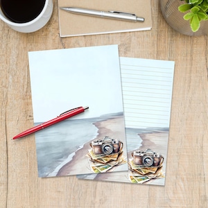 May include: Two sheets of stationery paper featuring a watercolour illustration of a vintage camera resting on a stack of photographs at the beach. One sheet is blank, and the other has horizontal lines for writing. A red pen lies across the paper.
