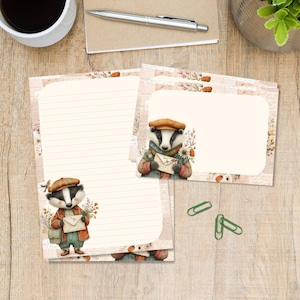 May include: Stationery set featuring a badger character wearing a cap and jacket, holding an envelope labelled Thank You. The set includes lined paper and a blank note card with floral accents, accompanied by two green paper clips on a wood surface.