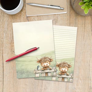 Puede incluir: Dos hojas de papel de carta con una ilustración de una vaca Highland marrón apoyada en una valla de madera en un campo. Una hoja está en blanco y la otra tiene líneas horizontales para escribir. Un bolígrafo rojo descansa sobre la hoja.